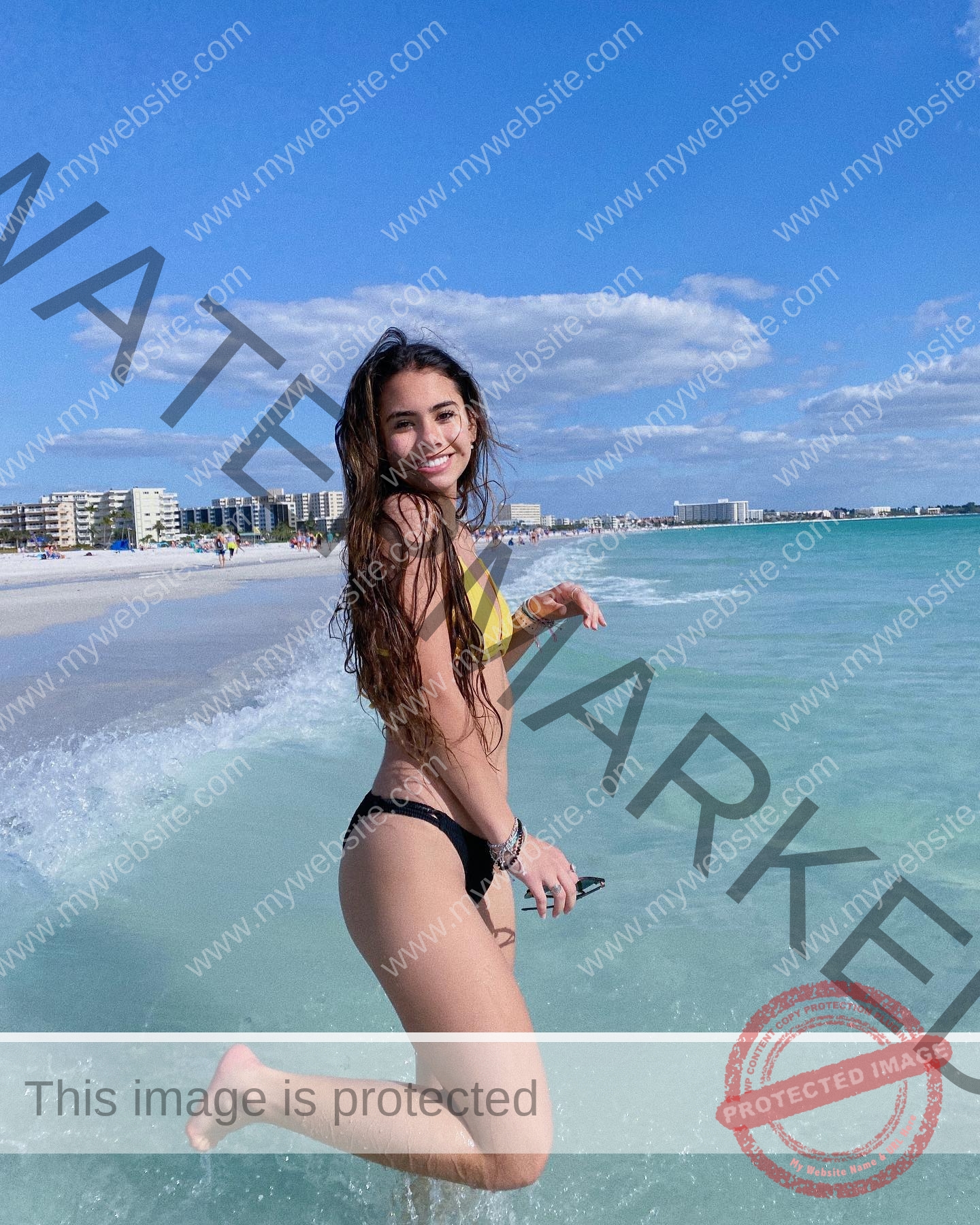 ciandra-carrico-trinity-ciandracarrico-4040 Ciandra Carrico, track athlete from Trinity, jumps playfully at the beach edge in a yellow bikini top with cityscape behind.