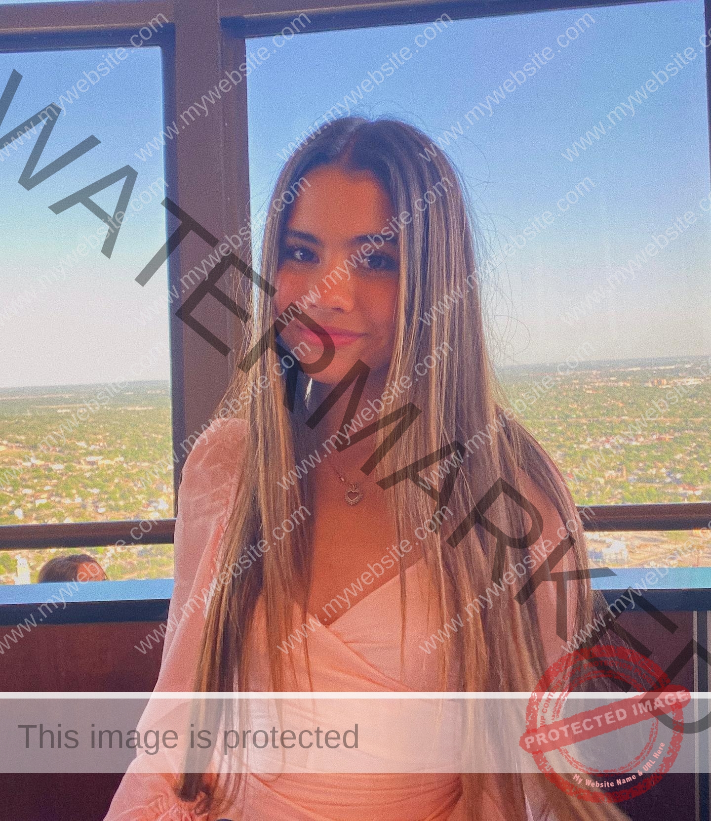 ciandra-carrico-trinity-ciandracarrico-4044 Ciandra Carrico, track athlete from Trinity, sits by a window in a pink dress and heart necklace; green landscape and blue sky behind.