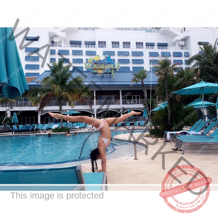ciandra-carrico-trinity-ciandracarrico-4050 Ciandra Carrico, track athlete from Trinity, does a handstand by a pool with lounge chairs, palms, and Margaritaville hotel behind.