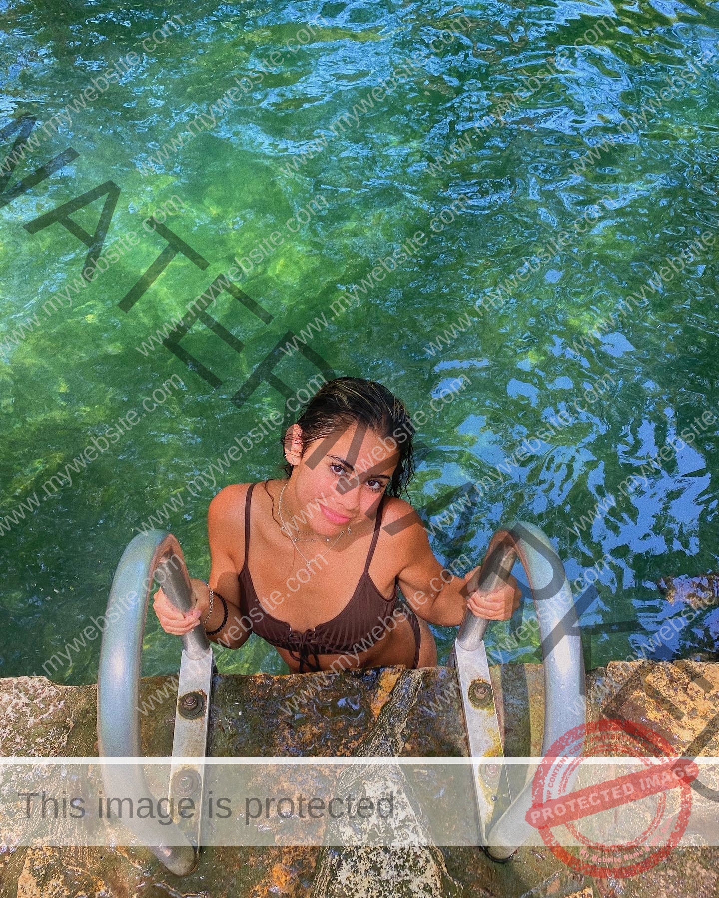 ciandra-carrico-trinity-ciandracarrico-4065 Ciandra Carrico, track athlete from Trinity, in a dark swimsuit smiles on a pool ladder, emerging from blue-green water in sunlight.