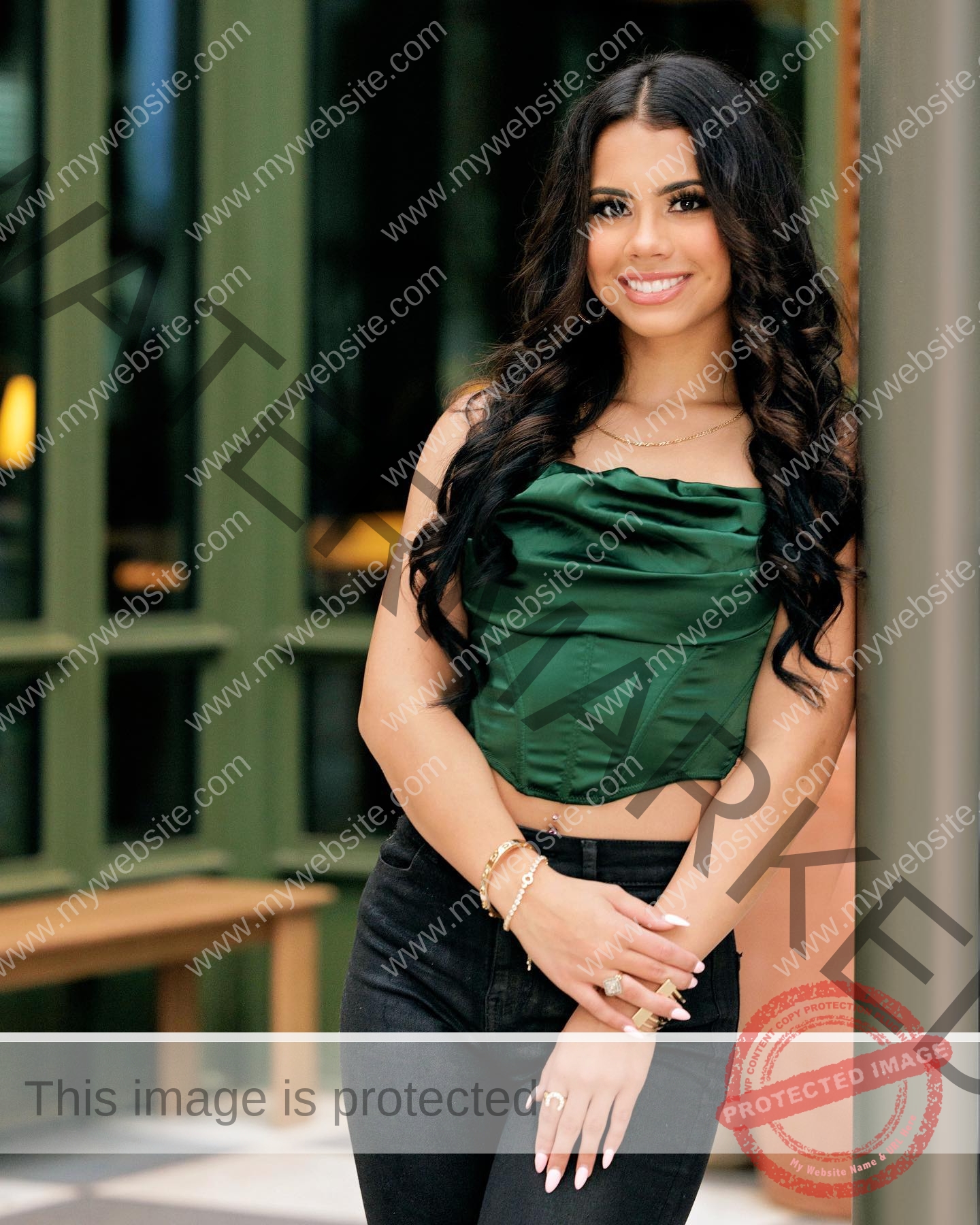 ciandra-carrico-trinity-ciandracarrico-4197 Ciandra Carrico, track athlete from Trinity, with long dark curly hair, smiles in a green top and black pants by a wall and green windows.