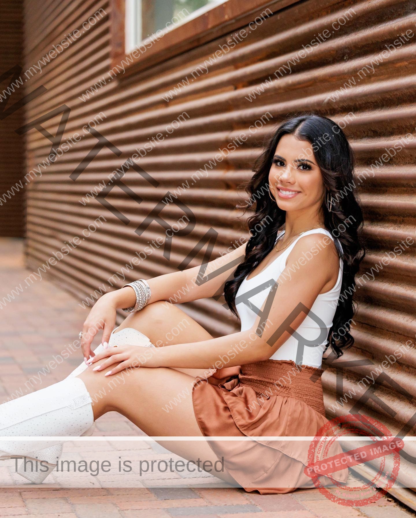 ciandra-carrico-trinity-ciandracarrico-4203 Ciandra Carrico, track athlete from Trinity, with long dark hair, sits on a brick walkway by metal wall, smiling at the camera.