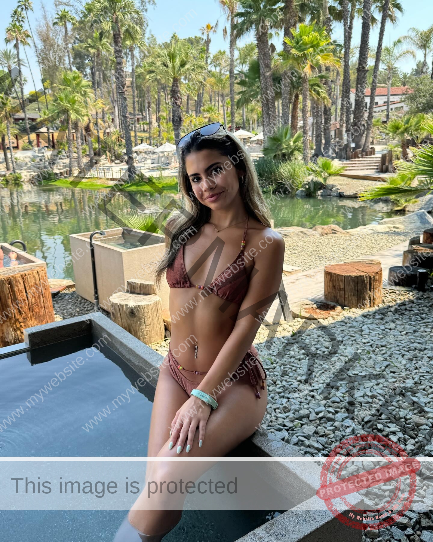 ciandra-carrico-trinity-ciandracarrico-4295 Ciandra Carrico, track athlete from Trinity, in a brown bikini sits poolside amid palm trees and a pond, smiling with sunglasses on her head.