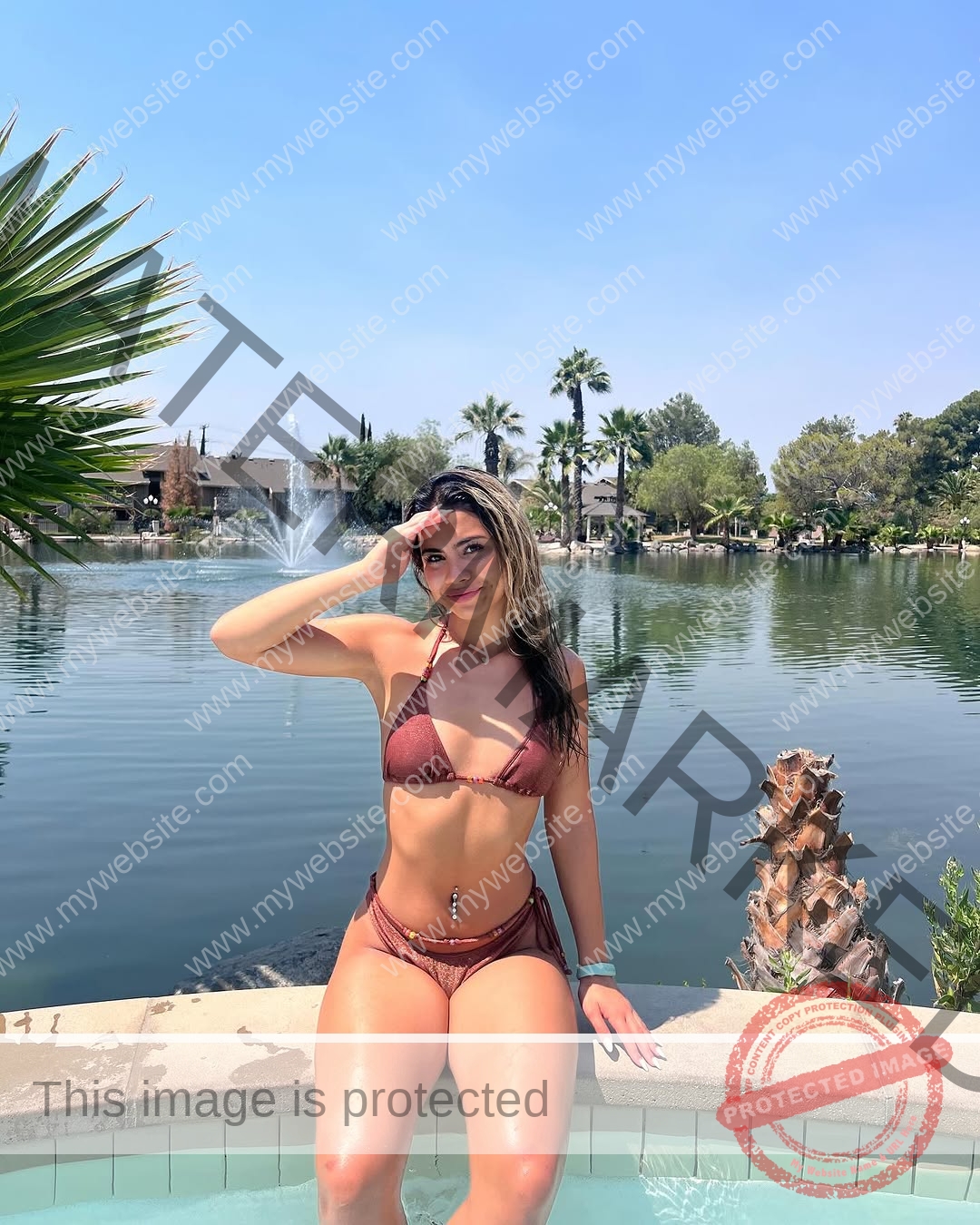 ciandra-carrico-trinity-ciandracarrico-4297 Ciandra Carrico, track athlete from Trinity, in a brown bikini sits poolside saluting; lake, fountain, palms, greenery behind.