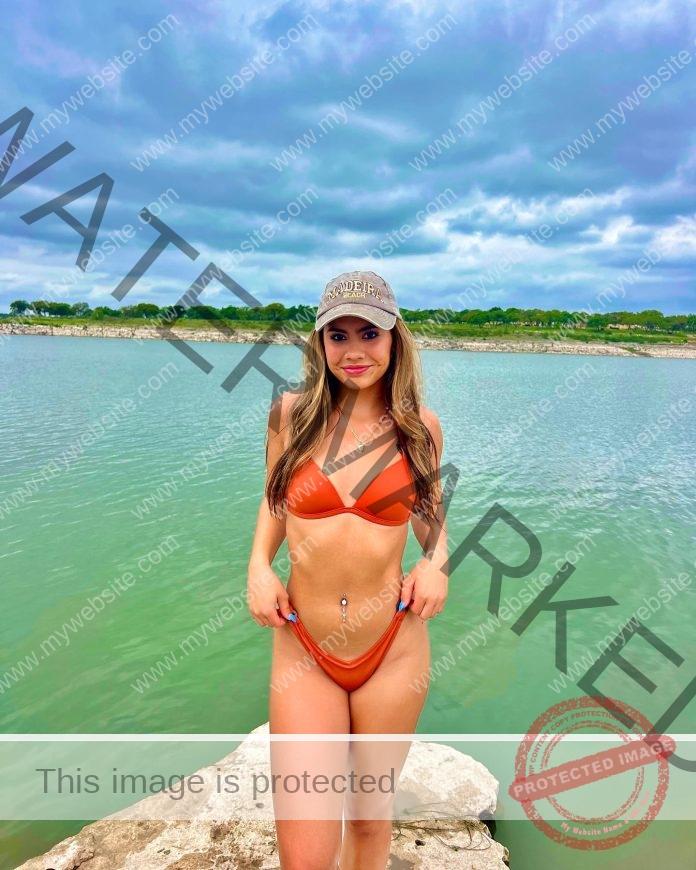 Ciandra Carrico, track athlete from Trinity, in an orange bikini and beige cap stands on a rock by a calm lake with greenery and clouds.