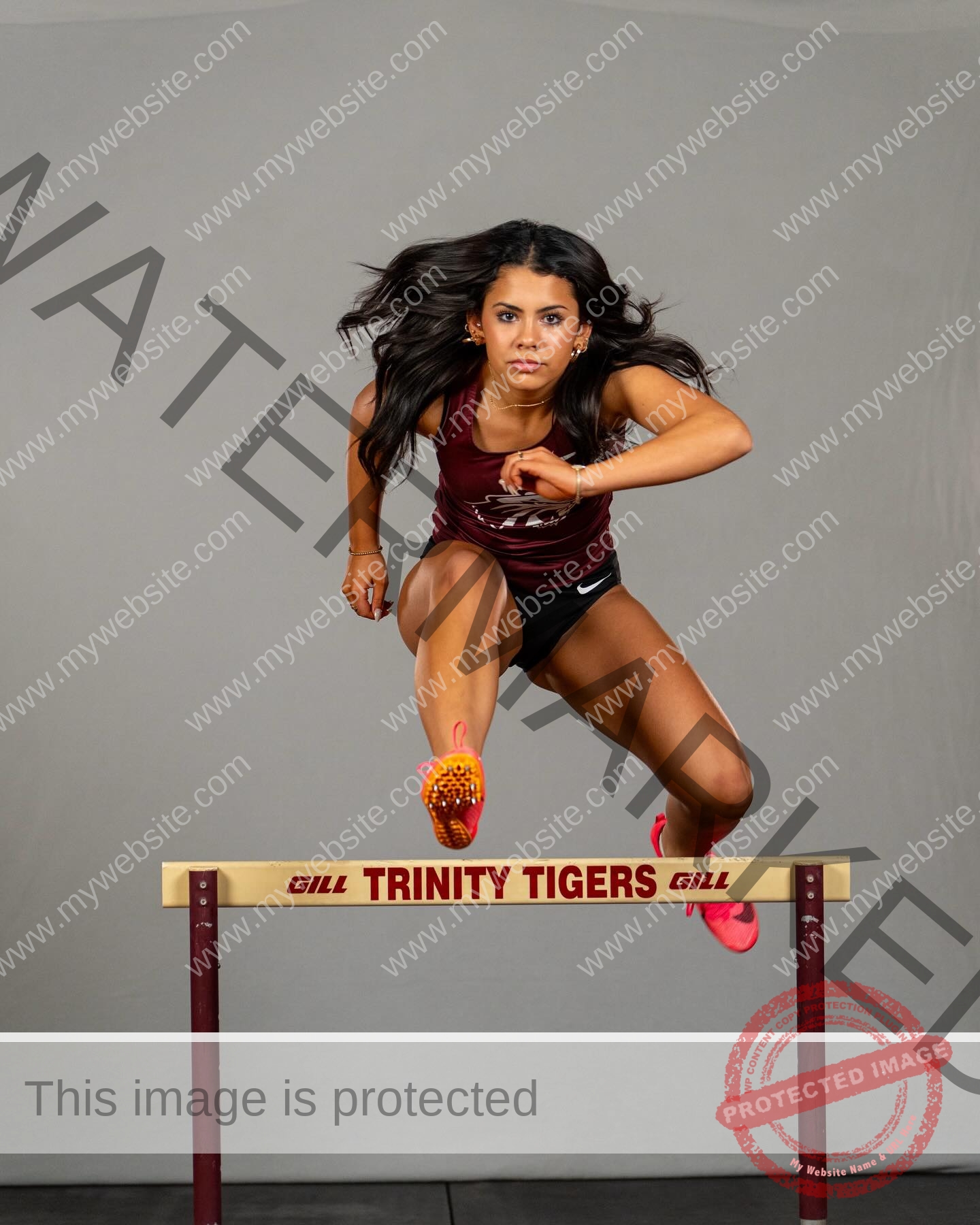 ciandra-carrico-trinity-ciandracarrico-4382 Ciandra Carrico, track athlete from Trinity, jumps mid-action over a TRINITY TIGERS hurdle in maroon and black with focused expression.