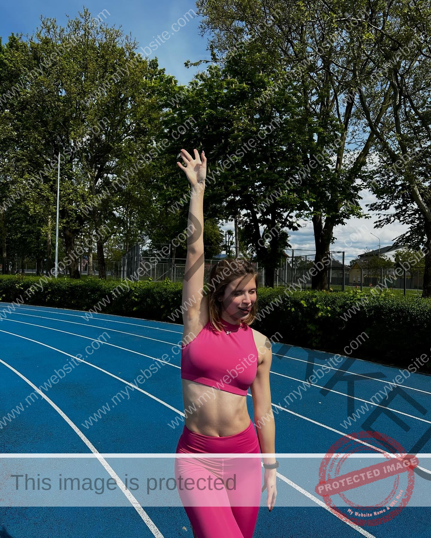 daria-polyanskaya-ukraine-polyanska.ya-x-08583 Daria-Polyanskaya, track athlete from Ukraine, in a bright pink outfit raises one arm on a blue running track amid green trees.