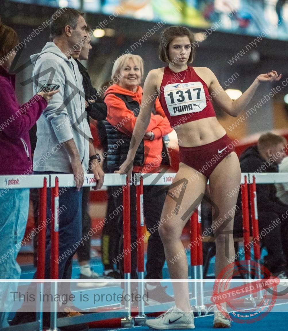daria-polyanskaya-ukraine-polyanska.ya-x-08637 Daria-Polyanskaya, track athlete from Ukraine, wears maroon sportswear and bib 1317 by indoor hurdles, gesturing as others watch.