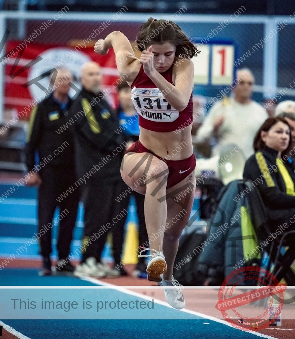 daria-polyanskaya-ukraine-polyanska.ya-x-08641 Daria-Polyanskaya, track athlete from Ukraine, in red with bib 131 jumps at an indoor meet; officials and spectators watch.