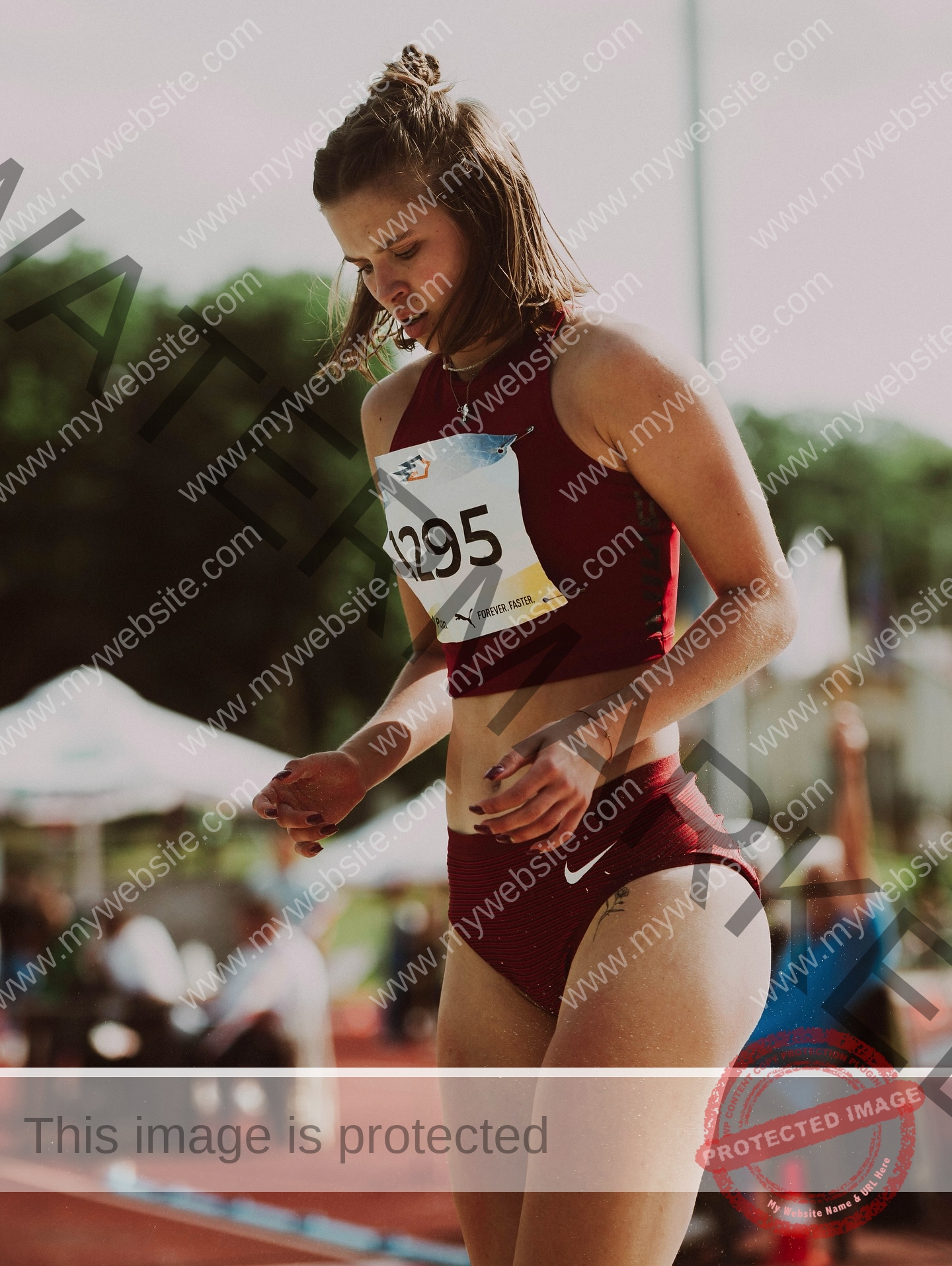 daria-polyanskaya-ukraine-polyanska.ya-x-08688 Daria-Polyanskaya, track athlete from Ukraine, in red uniform with bib 1295 stands focused on an outdoor track; blurred crowd and tents.