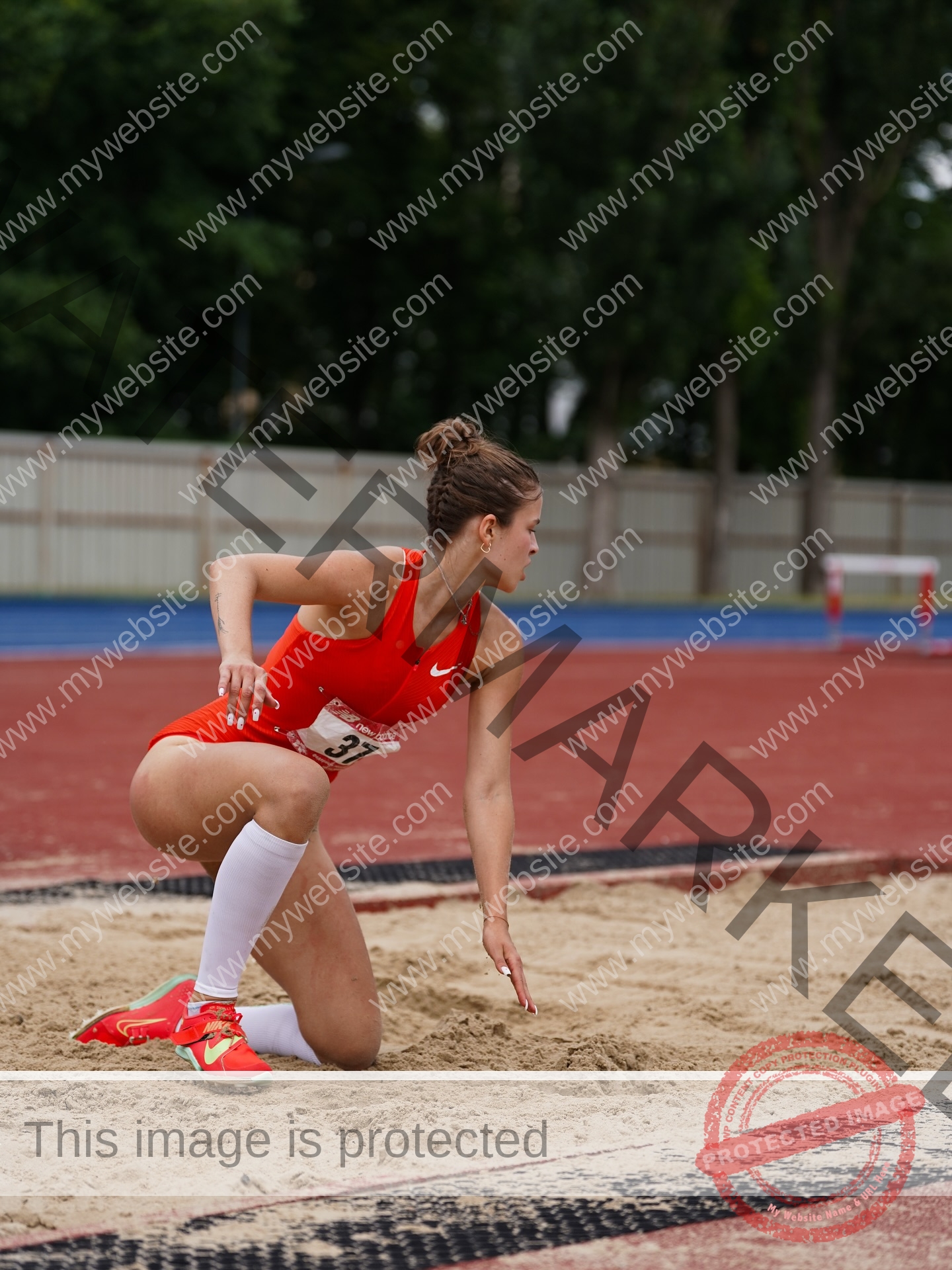 daria-polyanskaya-ukraine-polyanska.ya-x-08710 Daria-Polyanskaya, track athlete from Ukraine, in red uniform and white socks lands in sand during a long jump on an outdoor track.