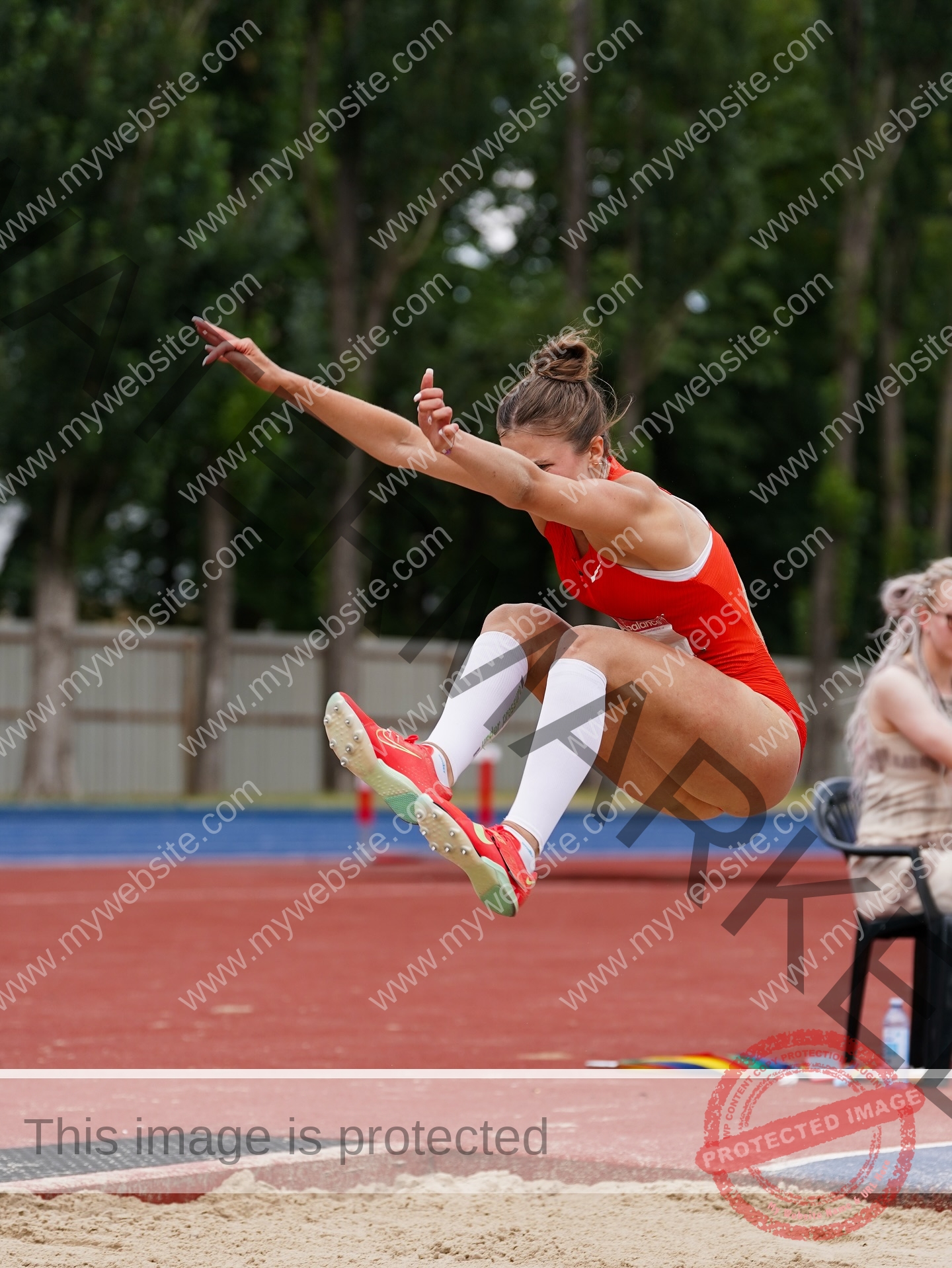 daria-polyanskaya-ukraine-polyanska.ya-x-08718 Daria-Polyanskaya, track athlete from Ukraine, in red uniform and white socks is airborne mid-long jump, ready to land in the sand.