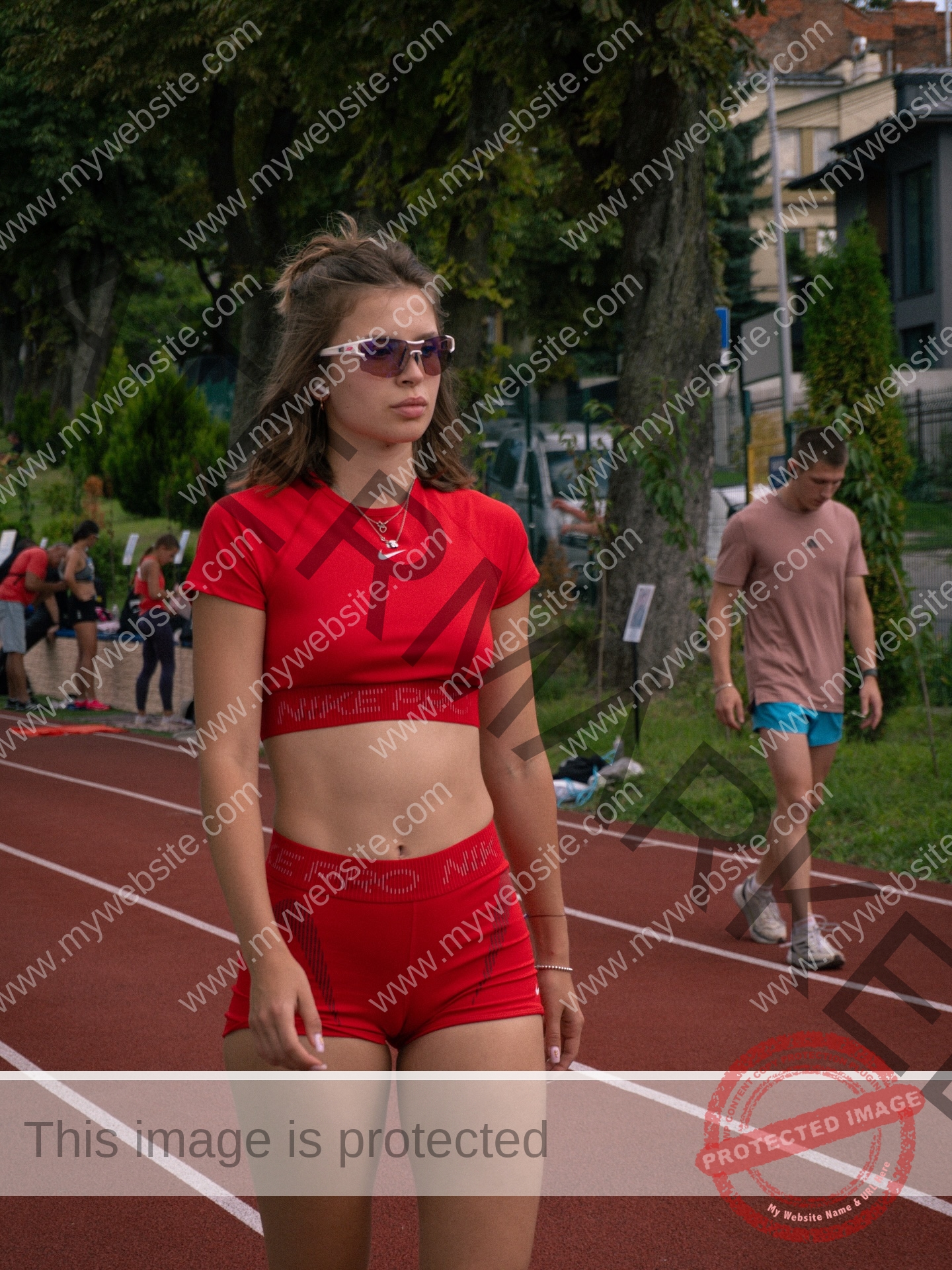 daria-polyanskaya-ukraine-polyanska.ya-x-08729 Daria-Polyanskaya, track athlete from Ukraine, in red athletic gear and sunglasses walks a track; man in pink behind her.