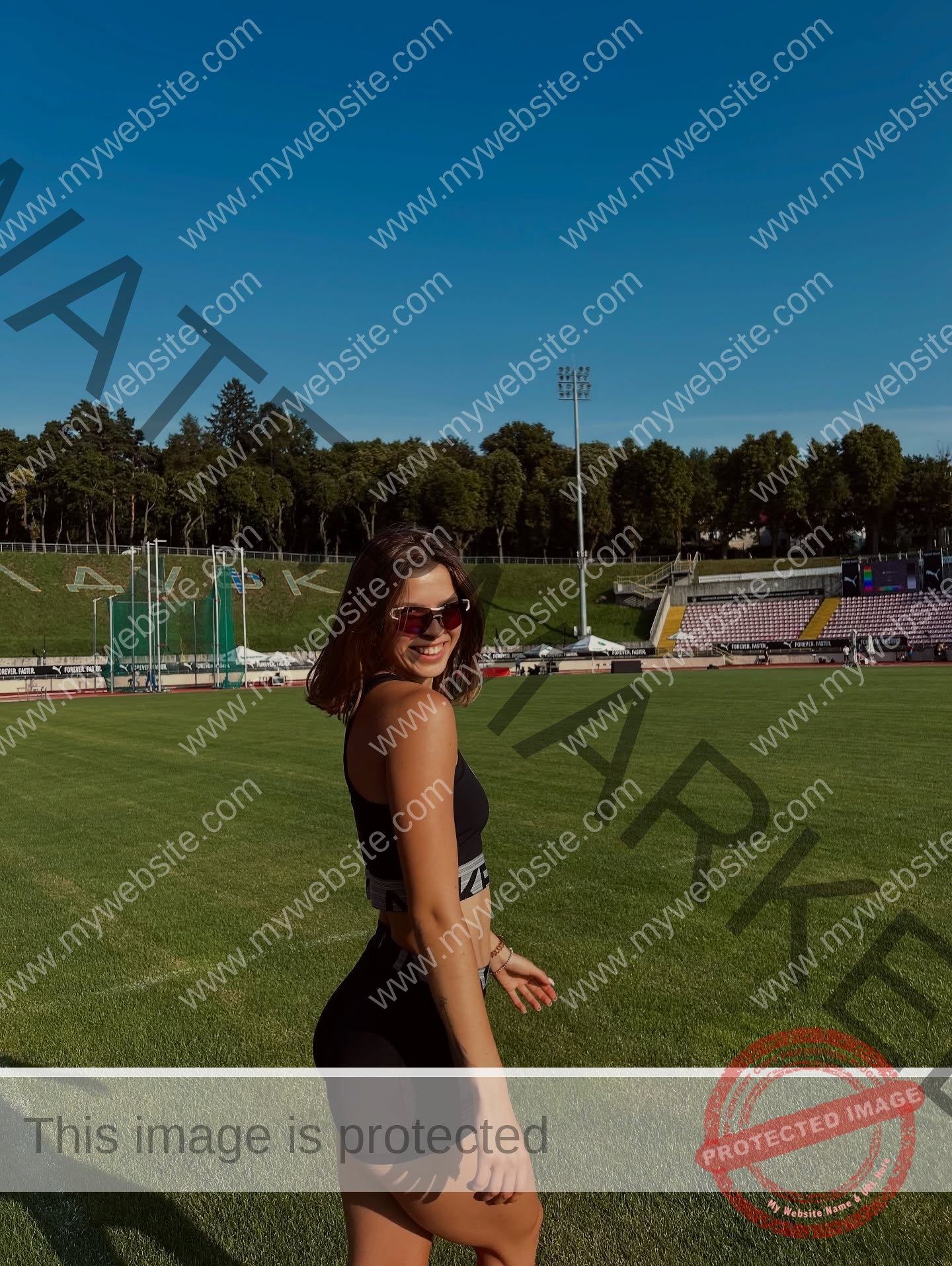 daria-polyanskaya-ukraine-polyanska.ya-x-08741 Daria-Polyanskaya, track athlete from Ukraine, wears sunglasses and athletic wear, smiling on a grassy sports field under a blue sky.