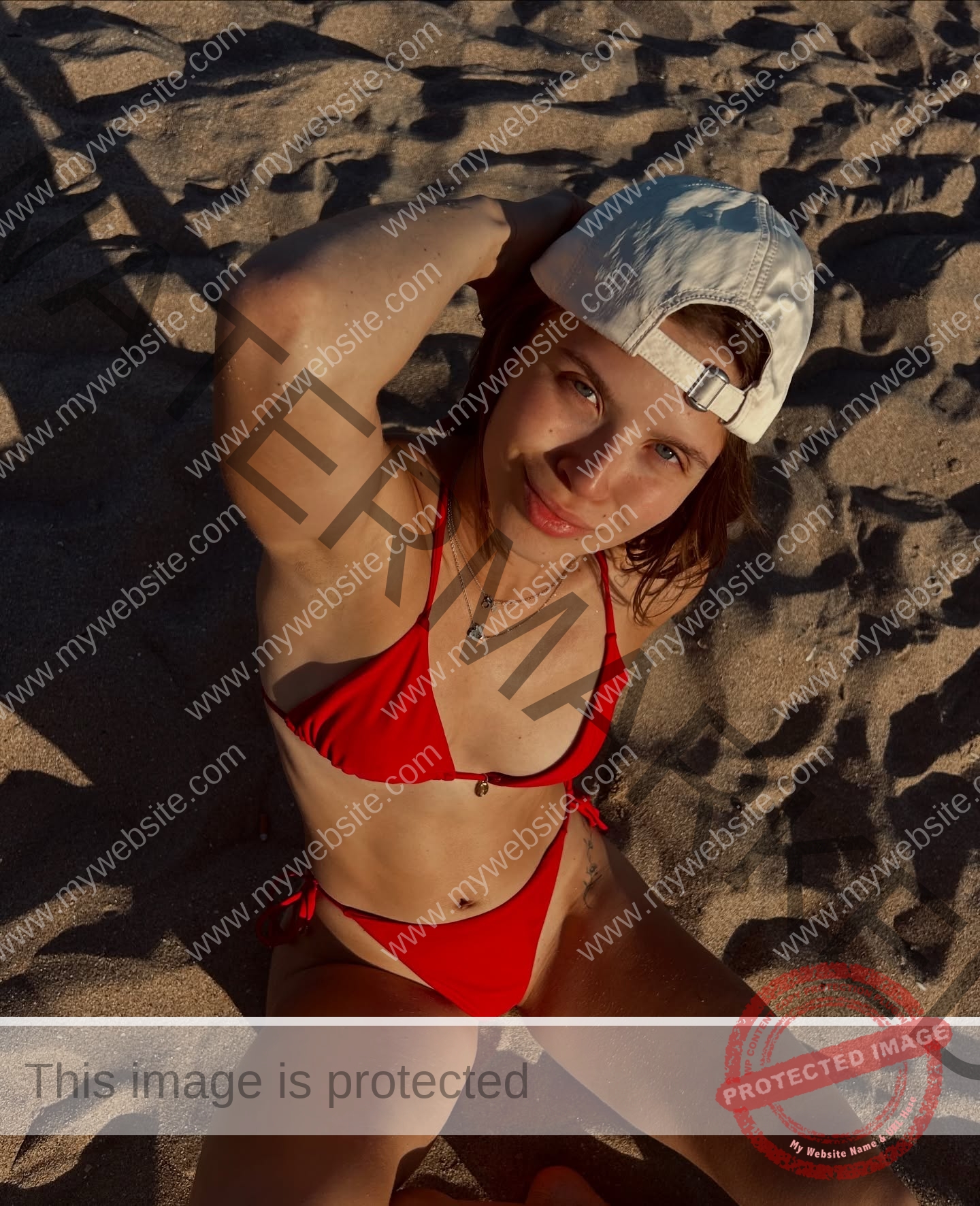 daria-polyanskaya-ukraine-polyanska.ya-x-08748 Daria-Polyanskaya, track athlete from Ukraine in red bikini and white cap sits on sandy beach, looking up at camera, sun casting shadows.