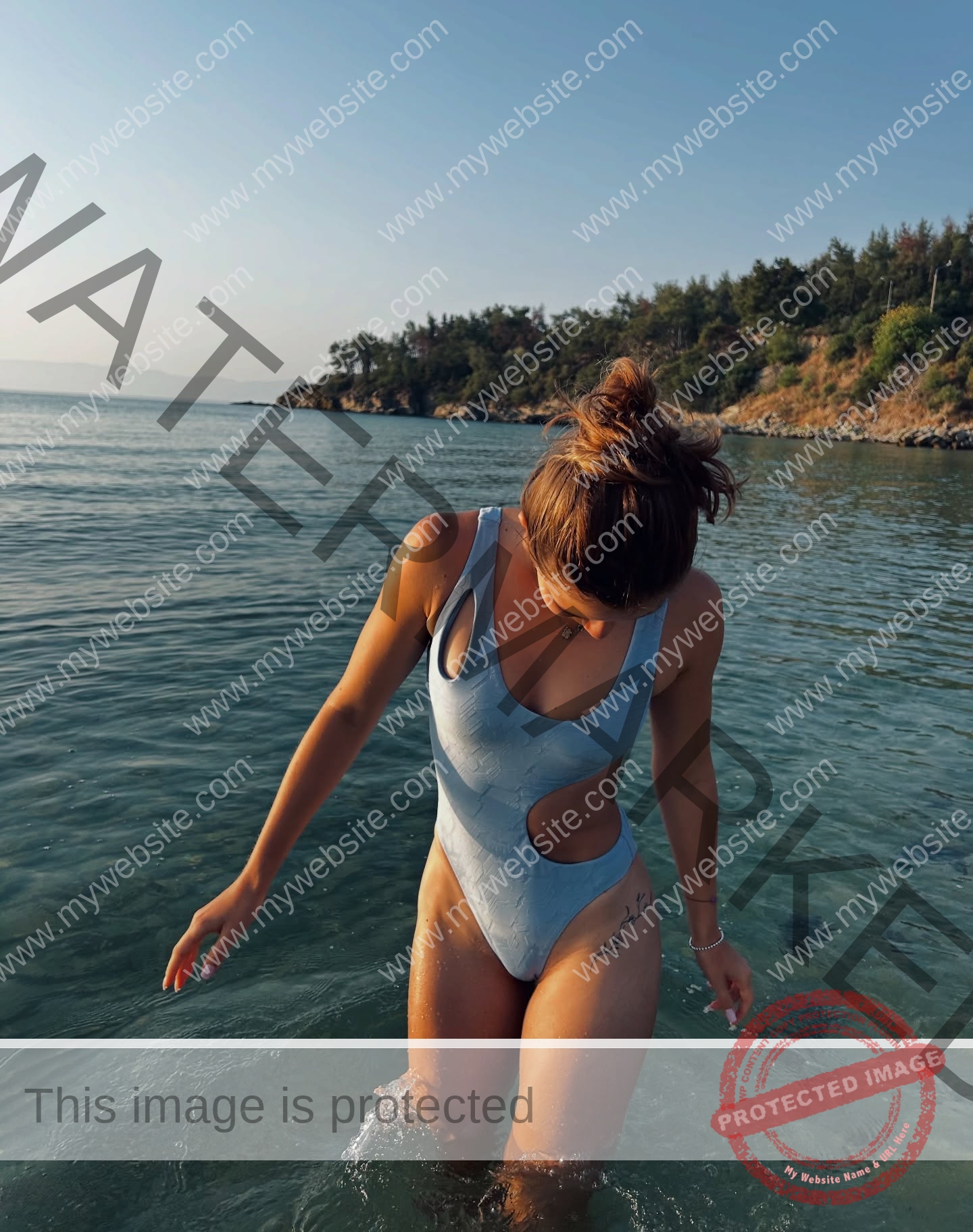 daria-polyanskaya-ukraine-polyanska.ya-x-08768 Daria-Polyanskaya, track athlete from Ukraine in a blue cut-out swimsuit stands in shallow water by a rocky shore, looking down.