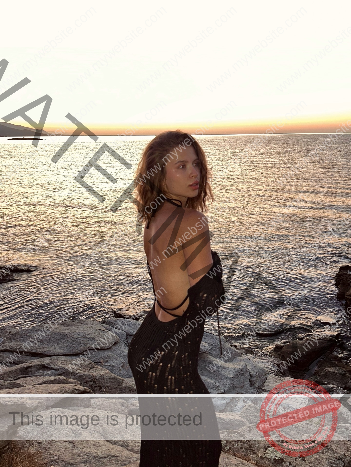 daria-polyanskaya-ukraine-polyanska.ya-x-08775 Daria-Polyanskaya, track athlete from Ukraine, in a black backless dress stands on rocky shore at sunset, golden sky behind her.