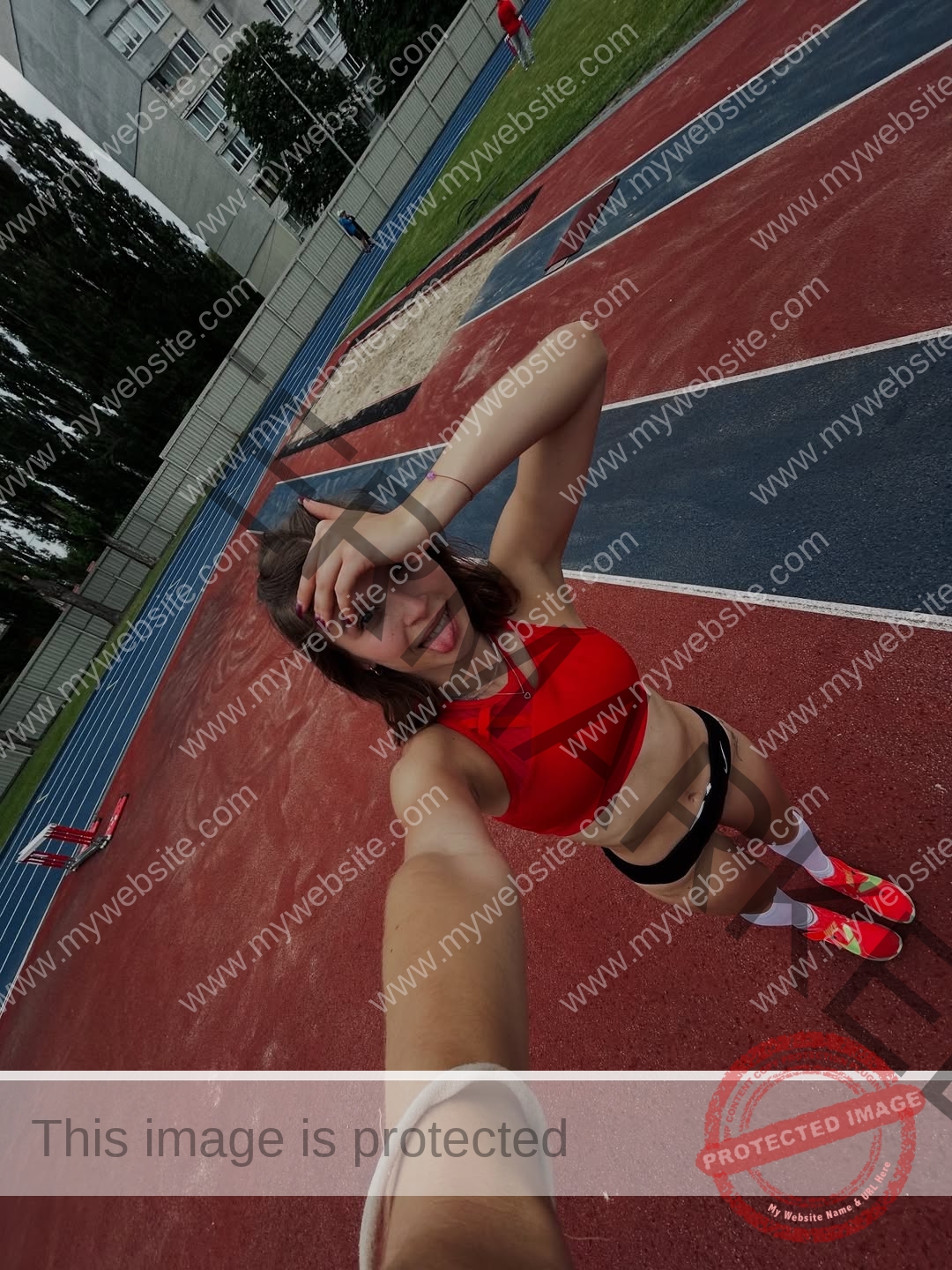 daria-polyanskaya-ukraine-polyanska.ya-x-09040 Daria-Polyanskaya, track athlete from Ukraine, takes a smiling selfie on an outdoor track by a sandpit with lanes and hurdles behind.