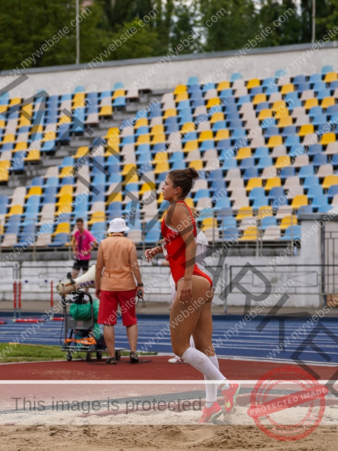 daria-polyanskaya-ukraine-polyanska.ya-x-09068 Daria-Polyanskaya, track athlete from Ukraine stands on a sandpit in a red outfit at a stadium with empty multicolored seats.