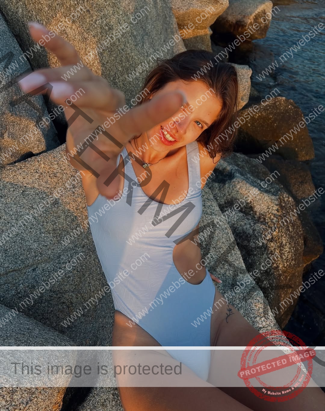 daria-polyanskaya-ukraine-polyanska.ya-x-09123 Daria-Polyanskaya, track athlete from Ukraine, sits on rocks by the water in a blue swimsuit, smiling and reaching to the camera.
