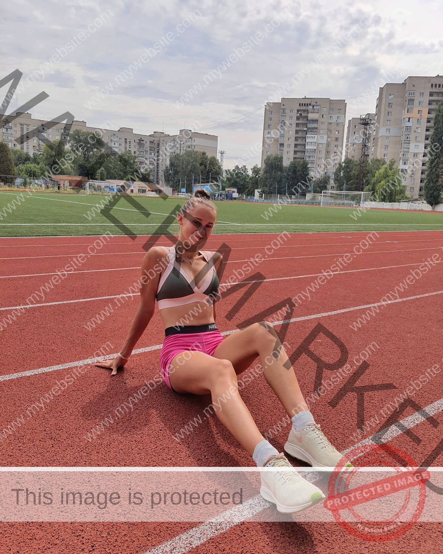 diana-honcharenka-ukraine-goncharenko_43-09538 Diana Honcharenka, track athlete from Ukraine sits on a red track in sports bra and pink shorts; tall buildings and greenery behind.