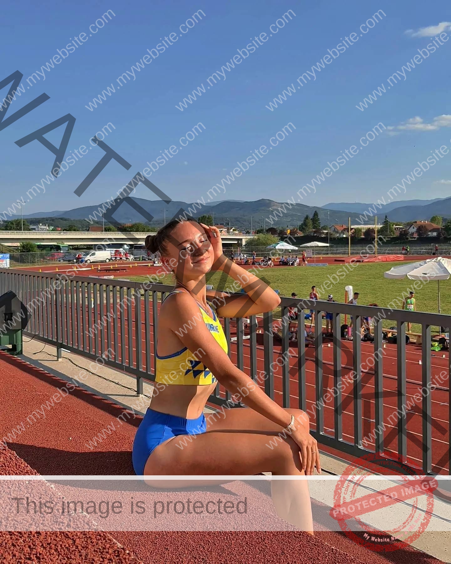 diana-honcharenka-ukraine-goncharenko_43-09544 Diana Honcharenka, track athlete from Ukraine, in yellow and blue gear sits smiling on a track, grassy field and mountains behind.