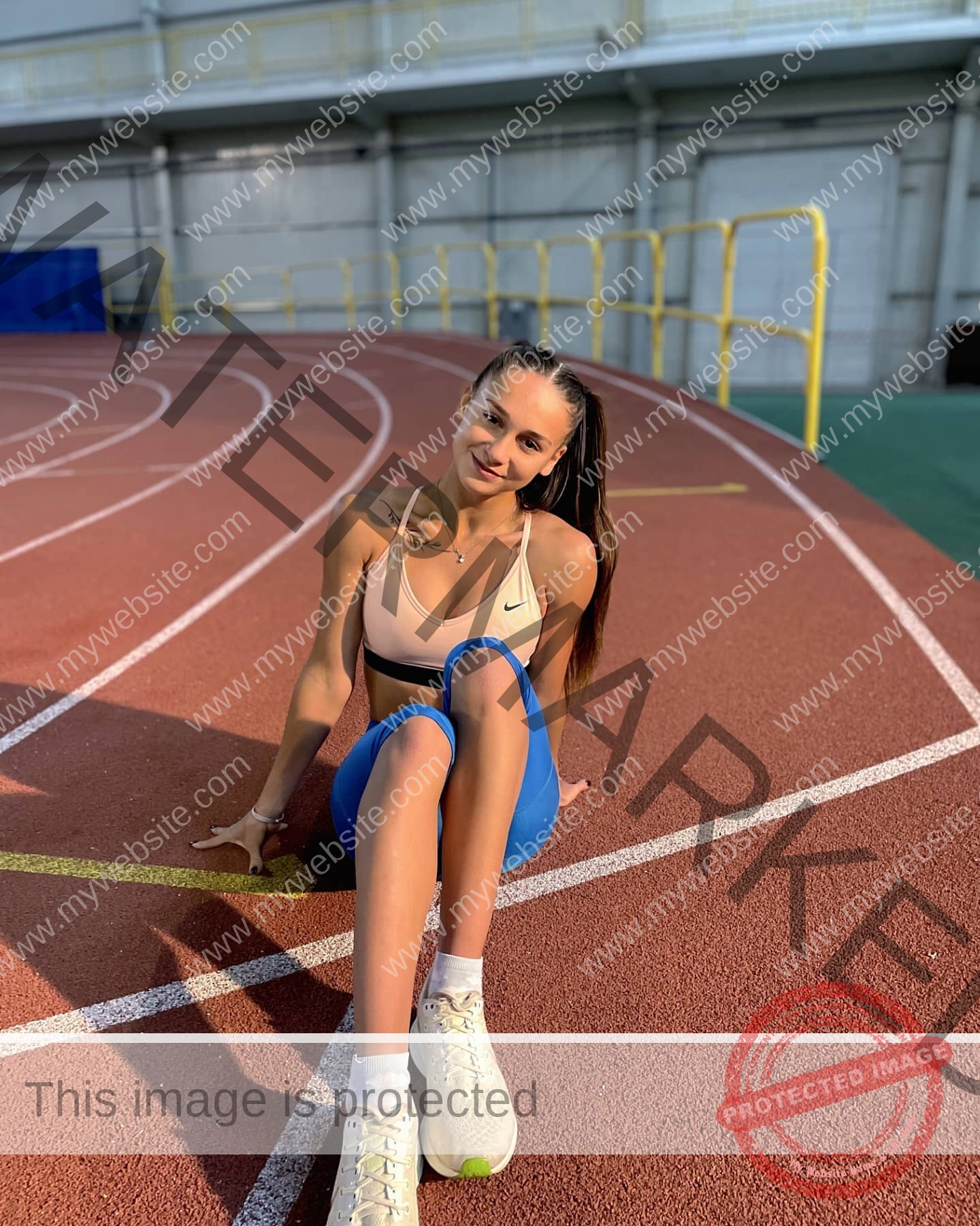 diana-honcharenka-ukraine-goncharenko_43-09587 Diana Honcharenka, track athlete from Ukraine, sits on an indoor track in blue leggings and white sneakers, smiling at the camera.