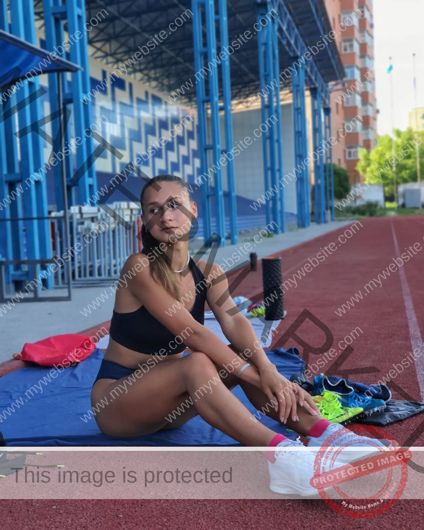 diana-honcharenka-ukraine-goncharenko_43-09627 Diana Honcharenka, track athlete from Ukraine, rests on a blue mat with running gear on a track; stadium and buildings behind.