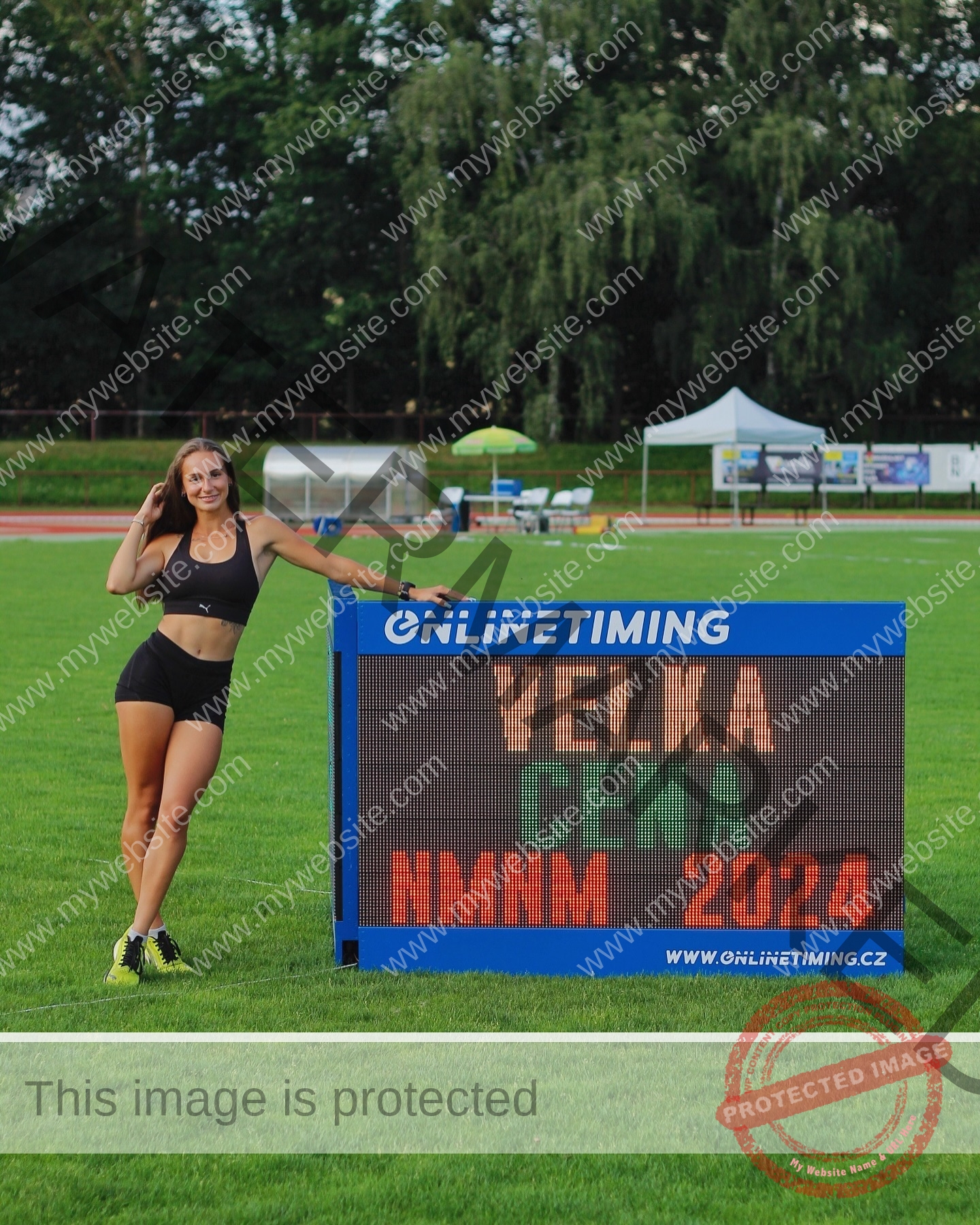 diana-honcharenka-ukraine-goncharenko_43-09832 Diana Honcharenka, track athlete from Ukraine, in sportswear poses by a VELKA CENA NMM 2024 sign on a field with tents behind.