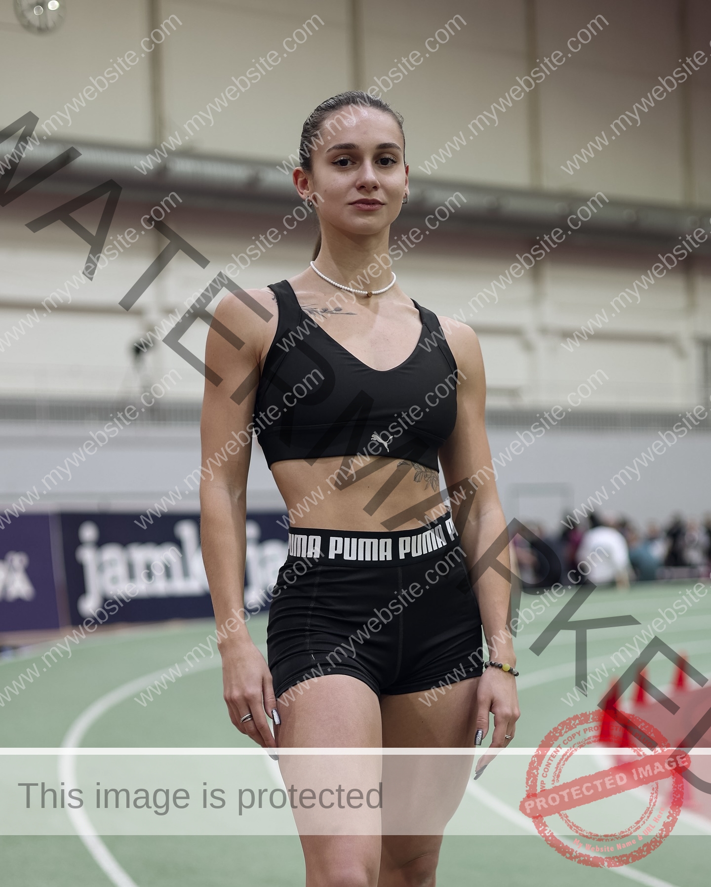 diana-honcharenka-ukraine-goncharenko_43-09869 Diana Honcharenka, track athlete from Ukraine, stands confidently on an indoor track in black sportswear, cones and spectators behind.