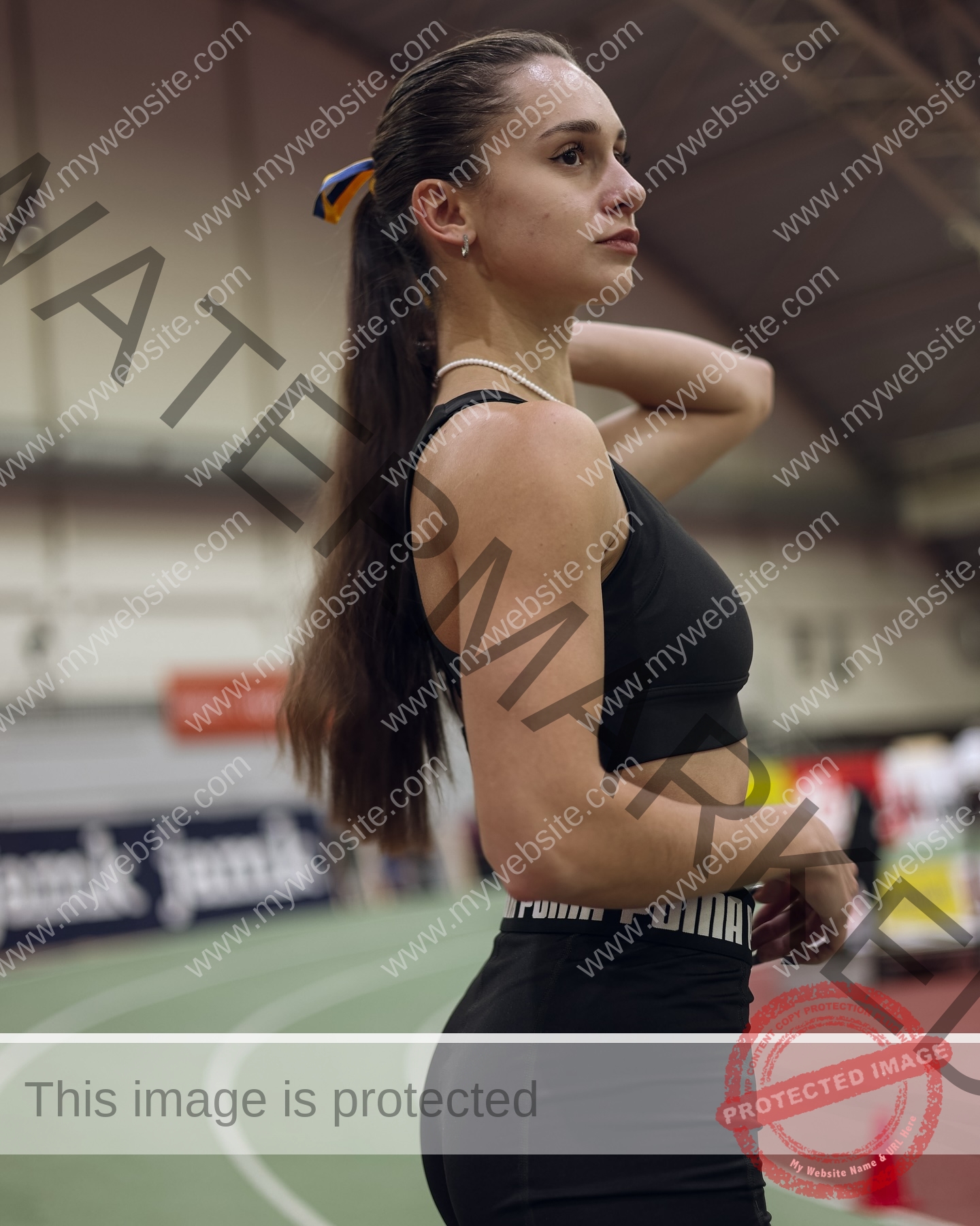 diana-honcharenka-ukraine-goncharenko_43-09871 Diana Honcharenka, track athlete from Ukraine, stands on an indoor track in black sportswear, focused and confident, with long hair tied back; stadium seats and banners are blurred in the background.