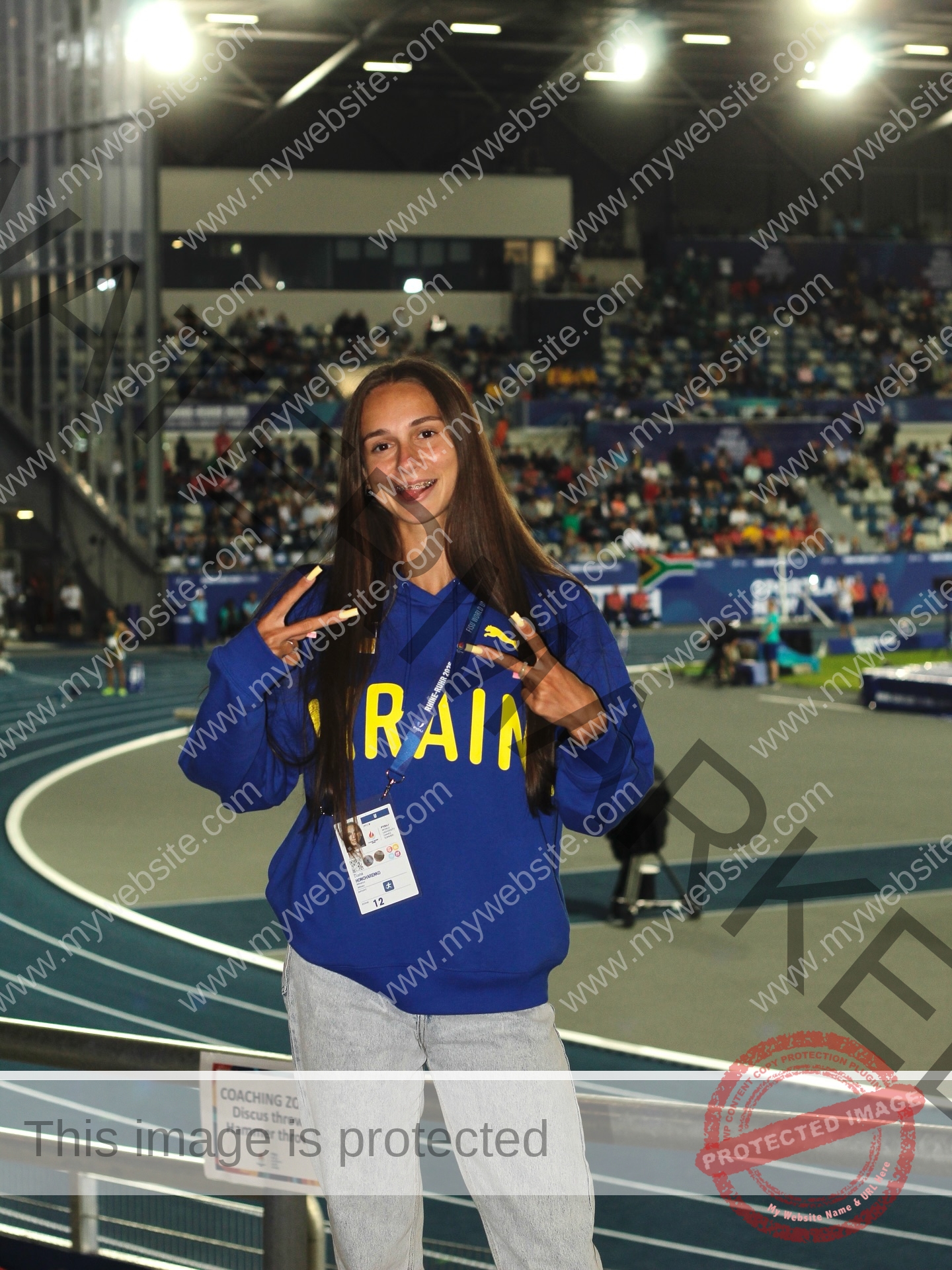 diana-honcharenka-ukraine-goncharenko_43-09933 Diana Honcharenka, track athlete from Ukraine, wears a blue hoodie and badge, smiling with peace signs before an indoor arena crowd.