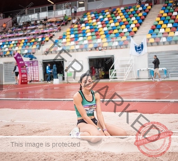 elena-corella-spain-elenacorella_-1052 Elena Corella A female track and field athlete in sportswear sits thoughtfully on a sandpit at a stadium with colorful empty seats nearby.