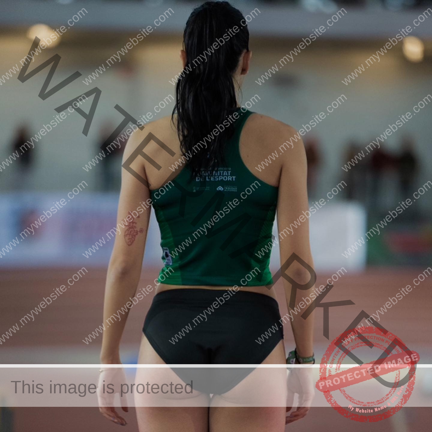 elena-corella-spain-elenacorella_-1055 Elena Corella A female track and field athlete stands on an indoor track, facing away, in a green top, black shorts, tattoo on left arm.