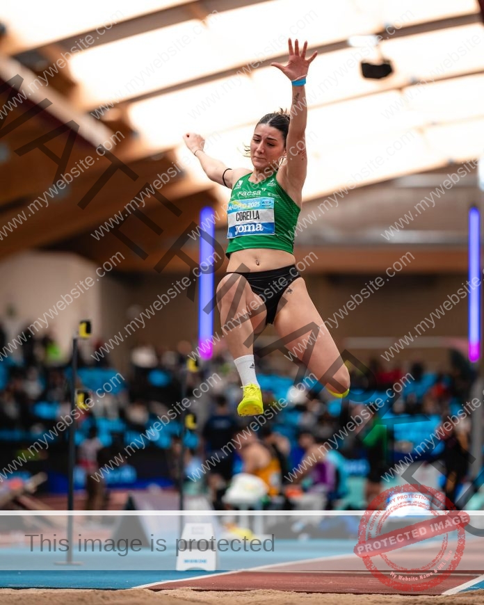 Elena Corella Spanish athlete Elena Corella soars mid-air during a long jump at an indoor track and field competition, with spectators and officials blurred in the background.