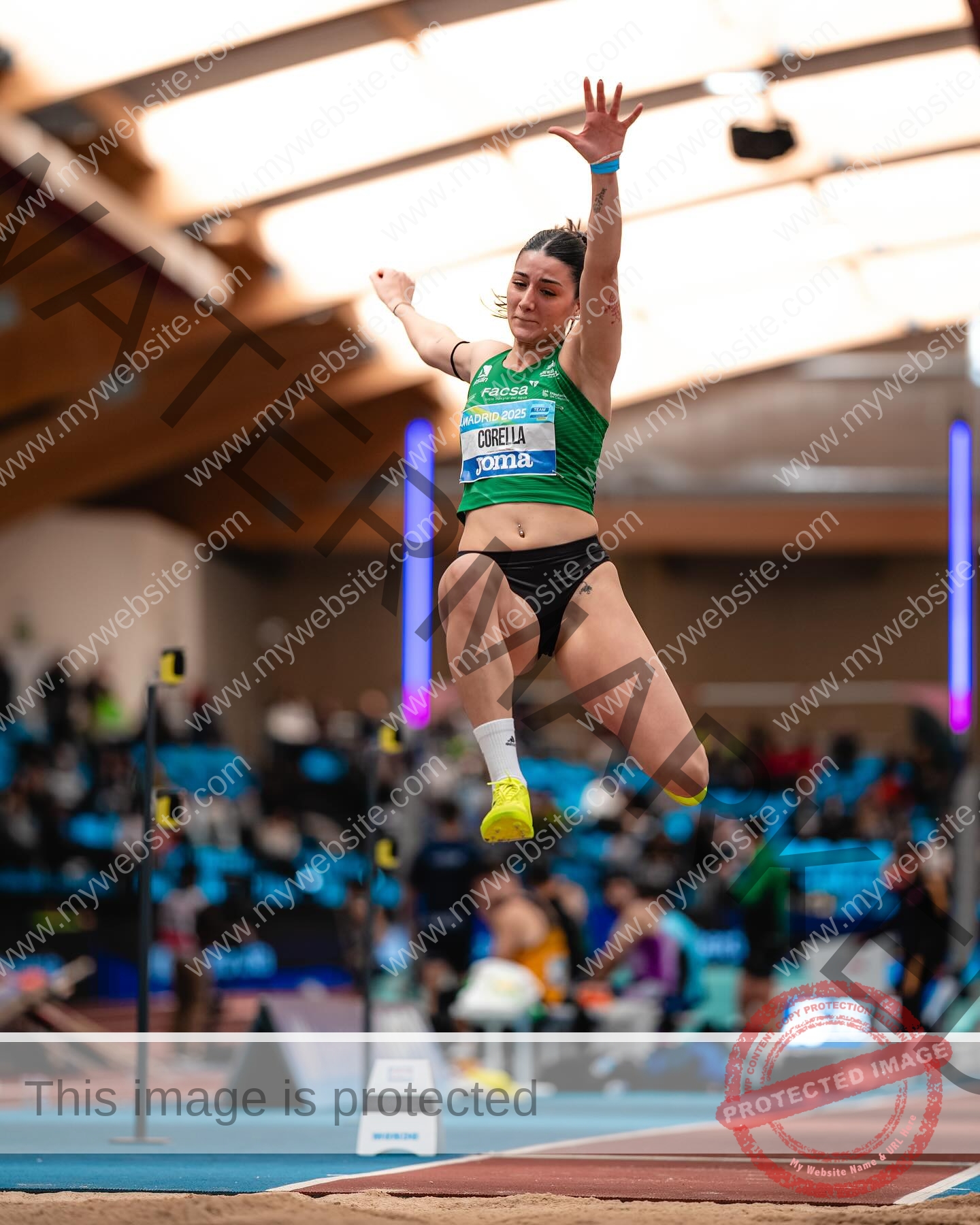 elena-corella-spain-elenacorella_-1076 Elena Corella Spanish athlete Elena Corella soars mid-air during a long jump at an indoor track and field competition, with spectators and officials blurred in the background.
