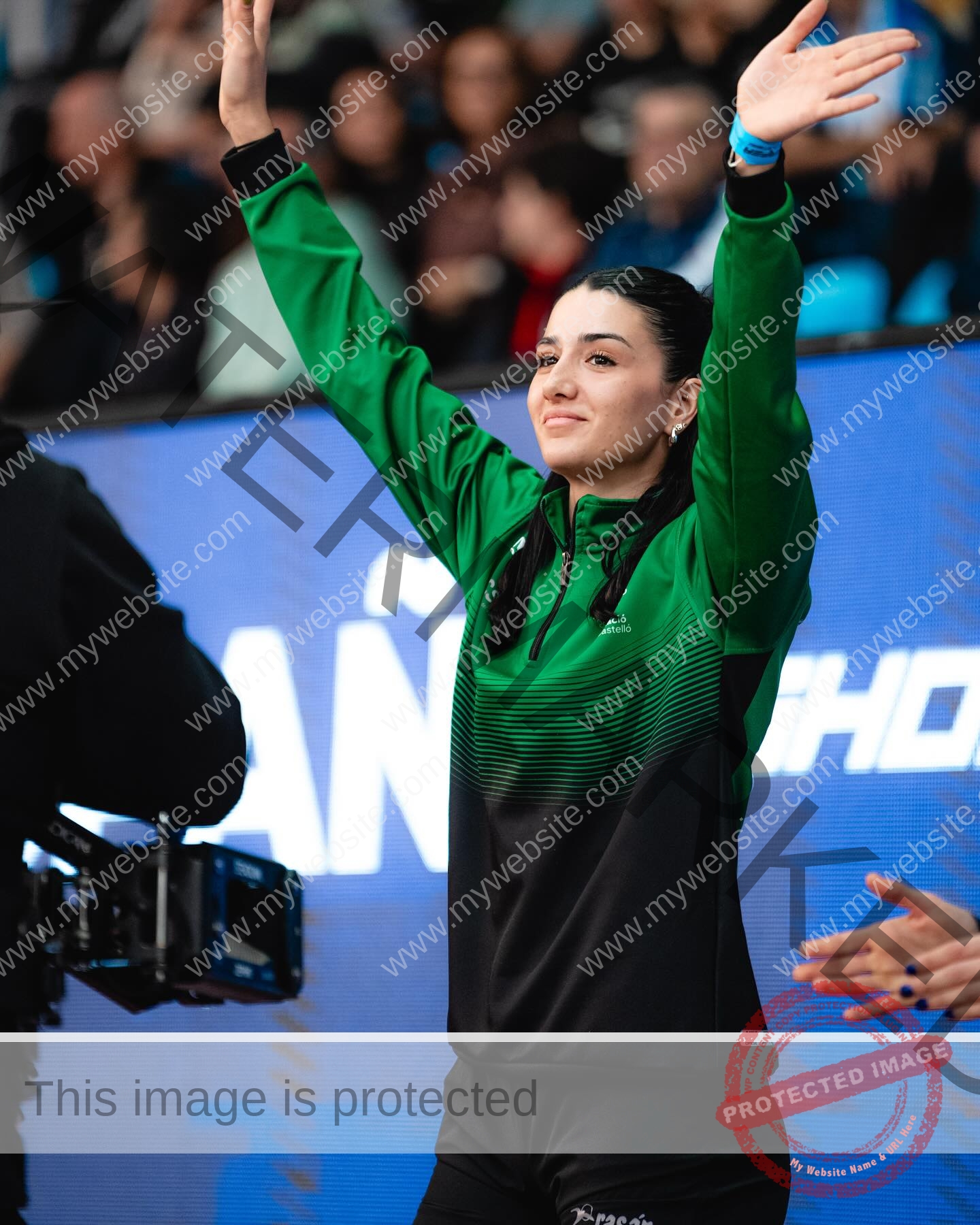 elena-corella-spain-elenacorella_-1077 Elena Corella A young woman, possibly track and field athlete Elena Corella, in a green and black sports jacket raises her arms and smiles, standing before a large screen and crowd at an indoor event in Spain.