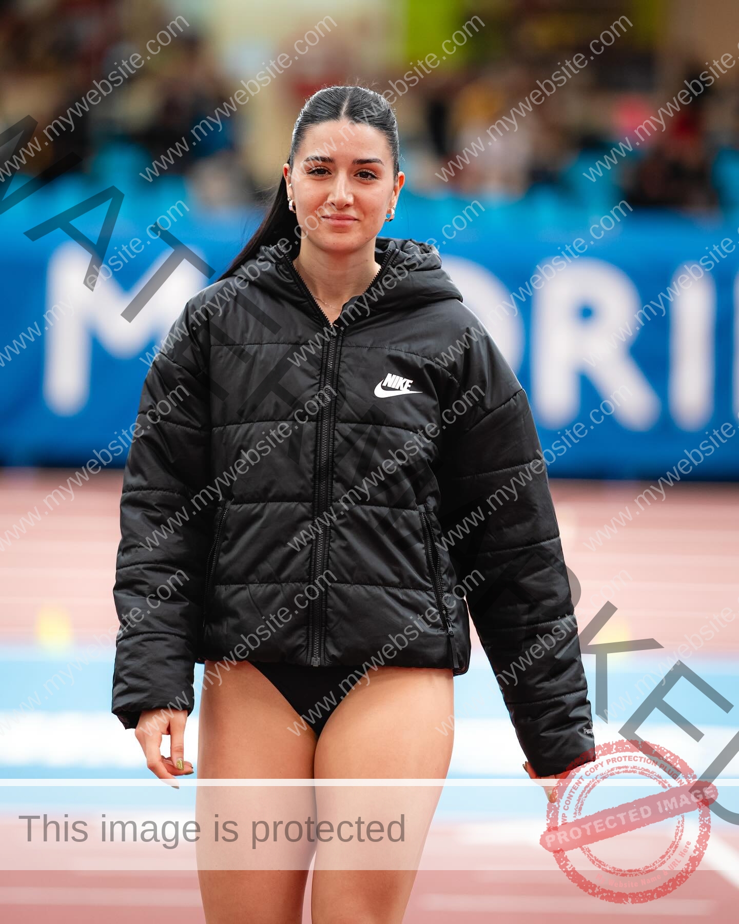 elena-corella-spain-elenacorella_-1078 Elena Corella Track and field athlete Elena Corella stands on an indoor track in Spain, wearing a black Nike jacket and shorts, with a blurred crowd and a blue MADRID sign in the background.