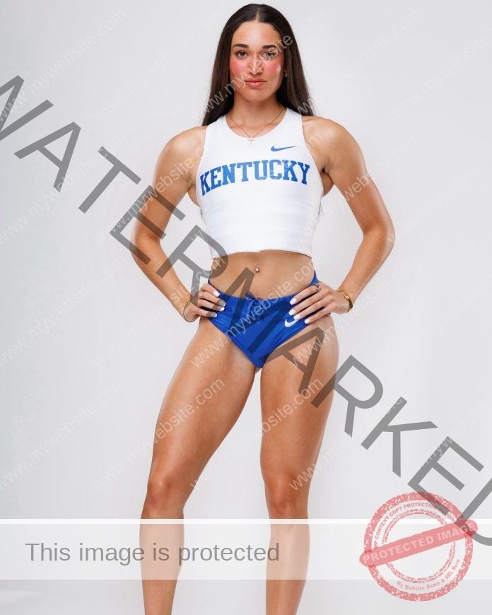Emmi Scales stands confidently against a white background in a KENTUCKY crop top and blue shorts, hands on hips, long dark hair.