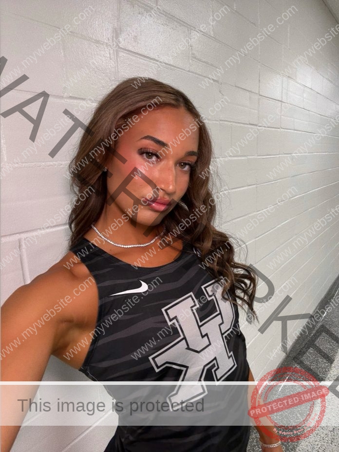 Emmi Scales, a woman with long wavy hair, takes a selfie in a hallway wearing a black H logo tank top for Kentucky track & field.