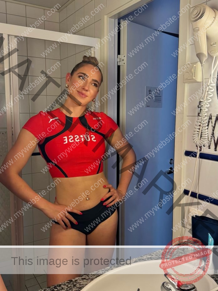 Giulia Wirth in a red SUISSE crop top and black underwear poses hands on hips, smiling in a bathroom with white tiles and hairdryer.