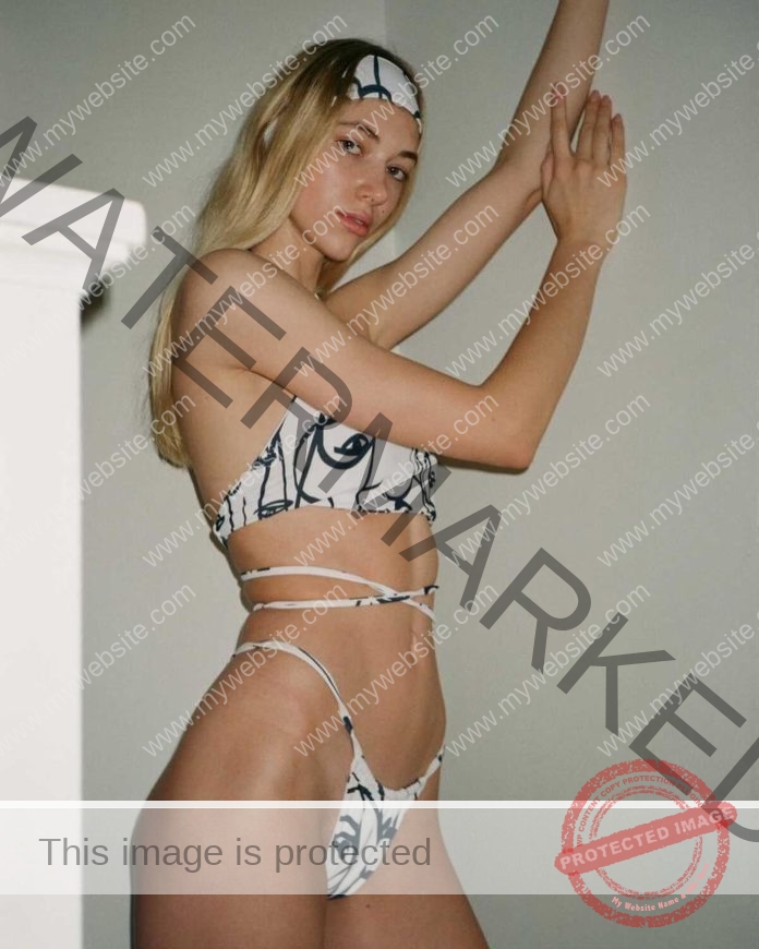 Grace Valette poses indoors in a white bikini with black line designs and a matching headband, confident against a plain wall.