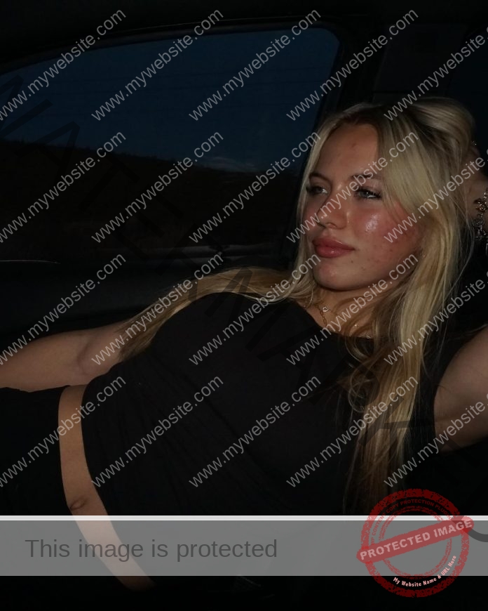 Hannah Bridge, a young woman with long blonde hair and a black crop top, reclines in a car seat at night in a CSUN track jacket.