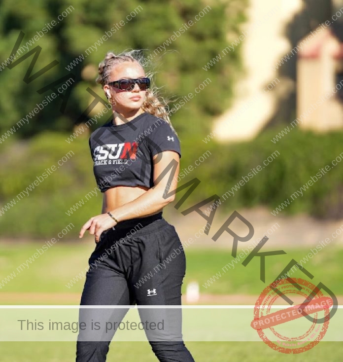 Hannah Bridge in sunglasses and a cropped black “Cal State Northridge Track & Field” shirt stands on a field, looking to the side.