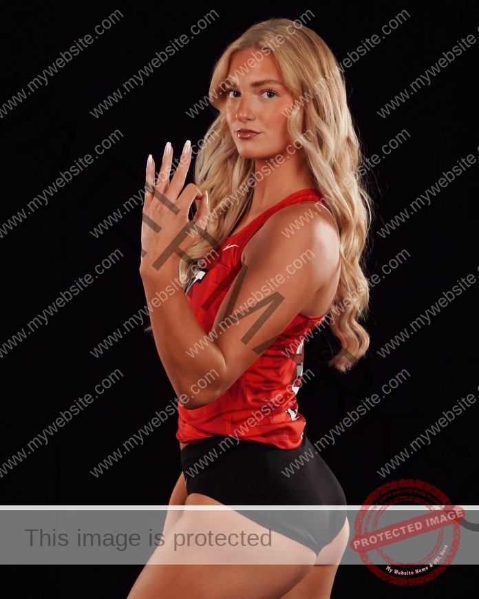 Kalli Knoble, a female volleyball player with long blonde hair in a red jersey and black shorts, poses against black holding up three fingers.