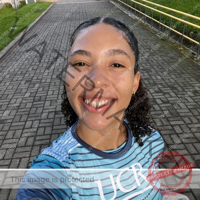 Kaylin Myrie, discus thrower from Costa Rica, a young woman with curly hair smiles on a path by grass and a yellow handrail.