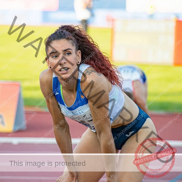 Laura Bueno A track and field athlete from Spain with long, curly red hair and a tattoo on her shoulder rests on the track, hands on knees, looking focused and slightly fatigued after a race.