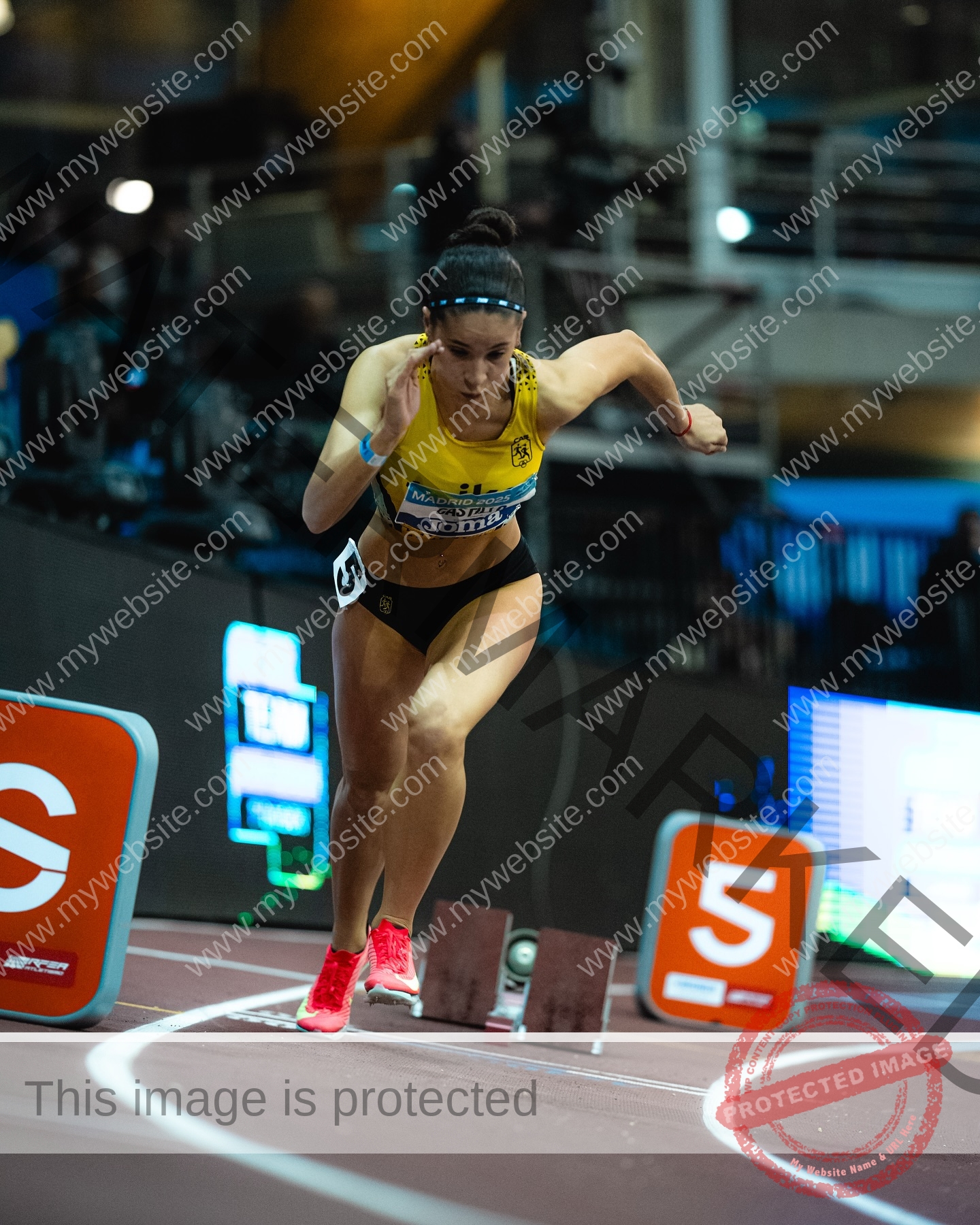 laura-castillo-spain-laura_c.i_-3277 Laura Castillo Spanish track and field athlete Laura Castillo sprints from starting blocks in yellow and black on an indoor track, lane markers and electronic screens visible.