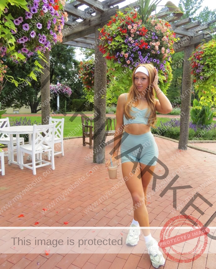 Madelyn Cisar Madelyn Cisar in a light blue workout outfit and white sneakers stands on a brick patio, holding a drink among flowers.