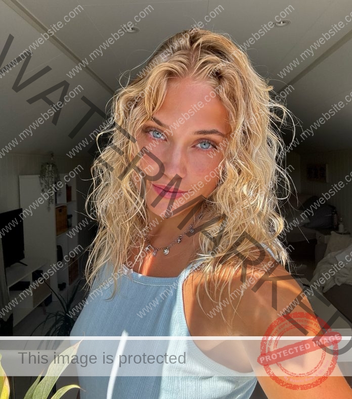 Malin Furuhaug Malin Furuhaug, with wavy blonde hair and blue eyes, takes a selfie in sunlight. Shelves, TV, and plants behind her.
